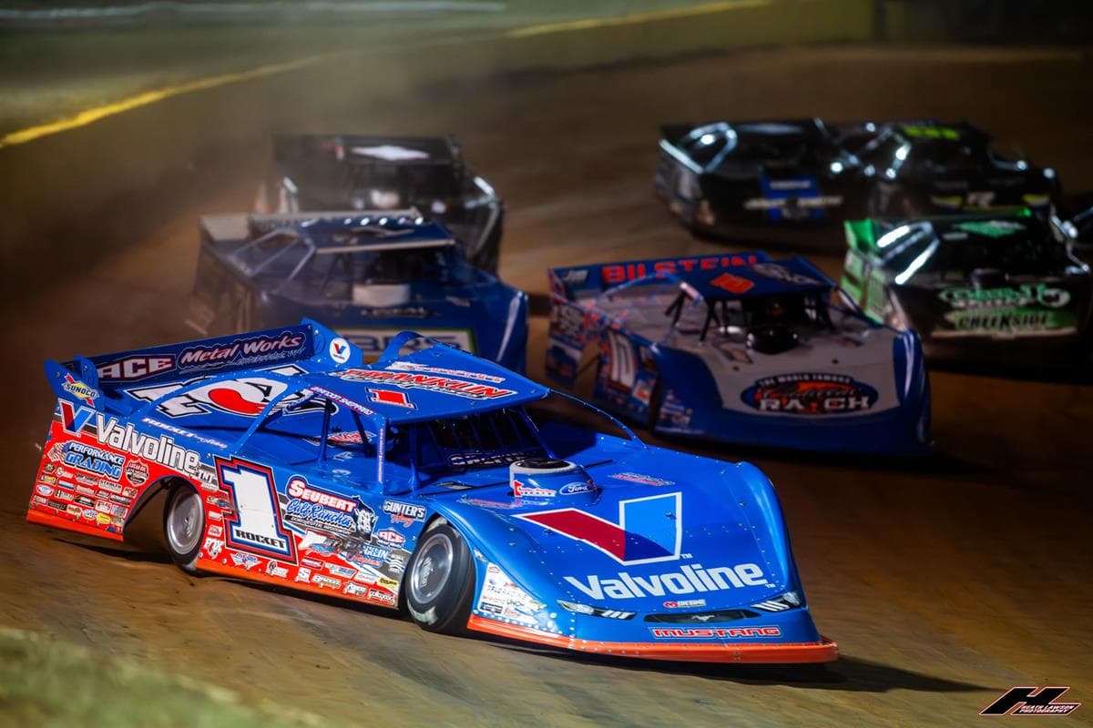 A Lucas Oil Late Model Dirt Series Super Late Model leads a pack of cars through a corner under the lights